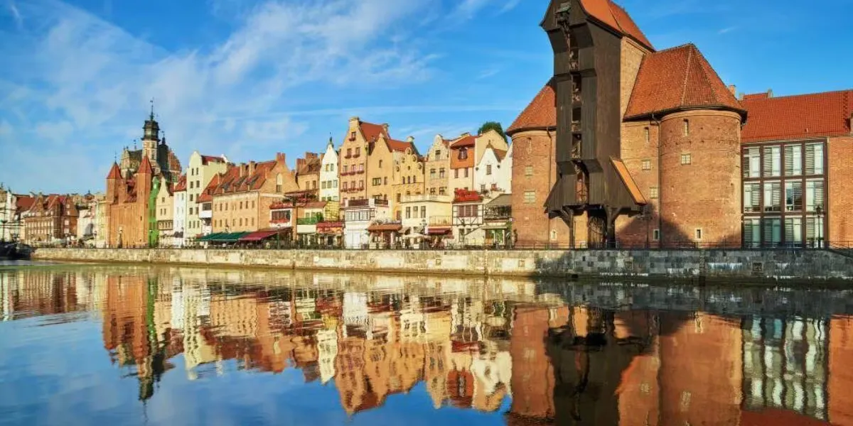 Sprzątanie Gdańsk, Gdynia, Sopot - Trójmiasto i okolice - Historyczne nabrzeże w Trójmieście ze starymi ceglanymi budynkami i charakterystycznym drewnianym dźwigiem odbijającym się w spokojnej rzece pod czystym, błękitnym niebem.