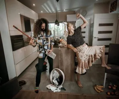 Sprzątanie Gdańsk, Gdynia, Sopot - Trójmiasto i okolice - Dwie osoby w kuchni udają, że grają muzykę - jedna uderza w miotłę jak w gitarę, a druga energicznie pozuje z uniesioną ręką. Pranie i jedzenie na podłodze sugerują sprzątanie po gościach po pracowitym trójmiejskim pobycie na wynajmie krótkoterminowym.